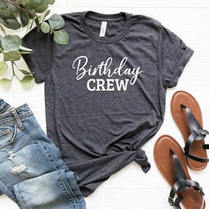 May include: Dark gray t-shirt with the words "Birthday CREW" in white script. The shirt is paired with blue jeans and brown sandals. The items are arranged on a white surface, suggesting a casual, everyday style.