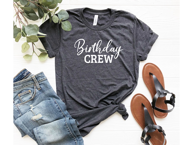 May include: Dark gray t-shirt with the words "Birthday CREW" in white script. The shirt is paired with blue jeans and brown sandals. The items are arranged on a white surface, suggesting a casual, everyday style.