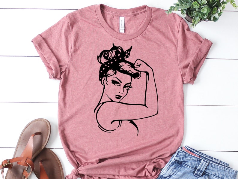Może przedstawiać: R&oacute;żowy T-shirt z czarną sylwetką kobiety z podniesioną pięścią. Kobieta ma w włosach bandanę w kropki.