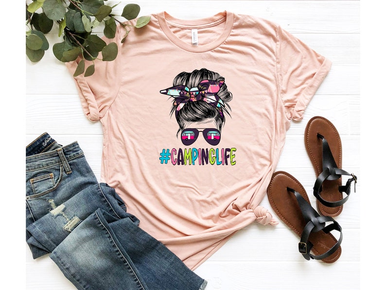 May include: A pink t-shirt with a graphic of a woman with a messy bun and sunglasses. The bun is decorated with a colorful floral pattern. The text "#CAMPLIFE" is printed in rainbow colors below the graphic.