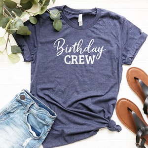 May include: A navy blue t-shirt with the words "Birthday CREW" in white script. The shirt is paired with denim shorts and brown sandals. Greenery is placed on the top left corner of the image.