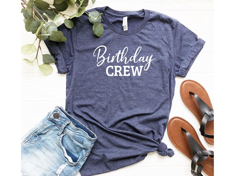 May include: A navy blue t-shirt with the words "Birthday CREW" in white script. The shirt is paired with denim shorts and brown sandals. Greenery is placed on the top left corner of the image.