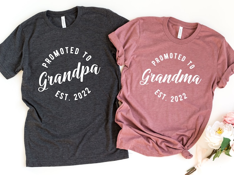 May include: Two t-shirts, one grey and one pink, with the text "Promoted to Grandpa Est. 2022" and "Promoted to Grandma Est. 2022" in white.