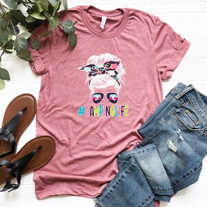 May include: A pink t-shirt with a graphic of a woman with a messy bun and sunglasses. The text "#CAMPINGLIFE" is printed below the graphic.
