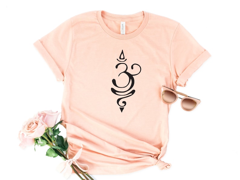 May include: A peach-colored t-shirt with a black Om symbol design. The shirt is laid flat on a white background with a pair of sunglasses and pink roses.