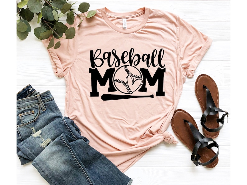 Op de afbeelding: Een roze T-shirt met de tekst "Baseball Mom" in zwarte letters. Een baseball met een hart erin is gecentreerd tussen de woorden "Mom". Een zwarte honkbalknuppel bevindt zich onder de baseball.