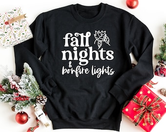 Może przedstawiać: Czarna bluza z białym napisem "fall nights & bonfire lights" i małym białym wzorem liścia.