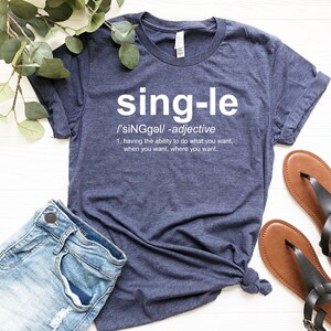May include: Navy blue t-shirt with the word "single" in white, along with its definition. The shirt is paired with denim shorts and brown sandals. Green leaves are visible in the background.