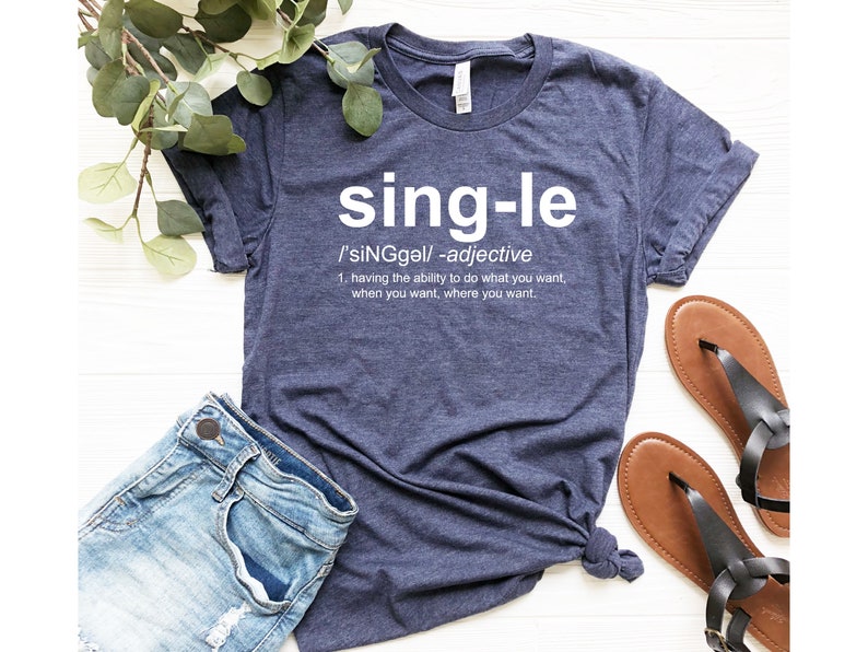 May include: Navy blue t-shirt with the word "single" in white, along with its definition. The shirt is paired with denim shorts and brown sandals. Green leaves are visible in the background.