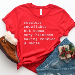May include: Red t-shirt with white text that reads "sweaters, snowflakes, hot cocoa, cozy blankets, baking cookies & santa". The shirt is laid out on a white surface with a pair of brown boots, blue jeans, and a small gift box.