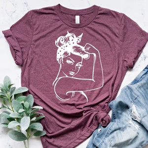 Może przedstawiać: Bordowy t-shirt z białym siluetem kobiety z podniesioną pięścią. Kobieta ma na sobie chustkę w kropki.
