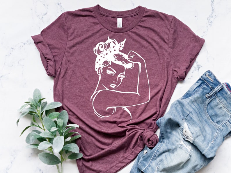Może przedstawiać: Bordowy t-shirt z białym siluetem kobiety z podniesioną pięścią. Kobieta ma na sobie chustkę w kropki.