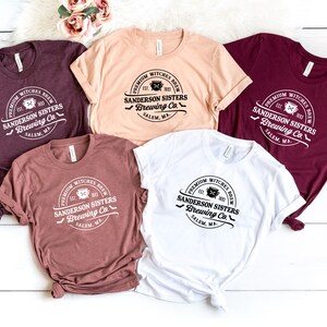 May include: Five different colored t-shirts with a white graphic print of a logo that says "Premium Witches Brew Est. 1963 Sanderson Sisters Brewing Co. Salem, MA."