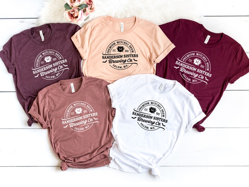 May include: Five different colored t-shirts with a white graphic print of a logo that says "Premium Witches Brew Est. 1963 Sanderson Sisters Brewing Co. Salem, MA."