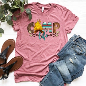 May include: A pink t-shirt featuring a graphic design of the word "camp" with a campfire and marshmallows. The text "life" is below the word "camp".