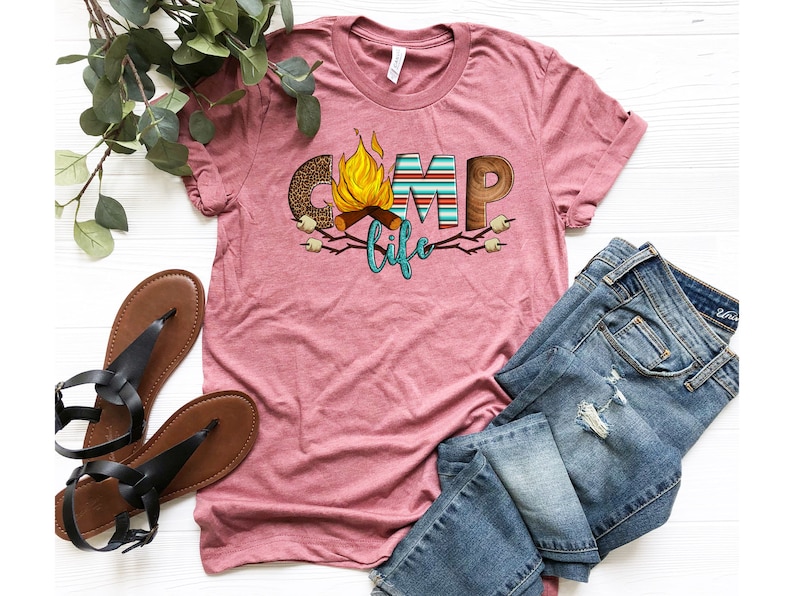 May include: A pink t-shirt featuring a graphic design of the word "camp" with a campfire and marshmallows. The text "life" is below the word "camp".