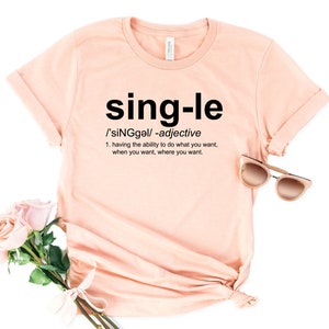 May include: A light peach-colored t-shirt with the word "single" in large black letters. Below the word is the definition of single as an adjective. A pair of sunglasses and a bouquet of pink roses are next to the shirt.
