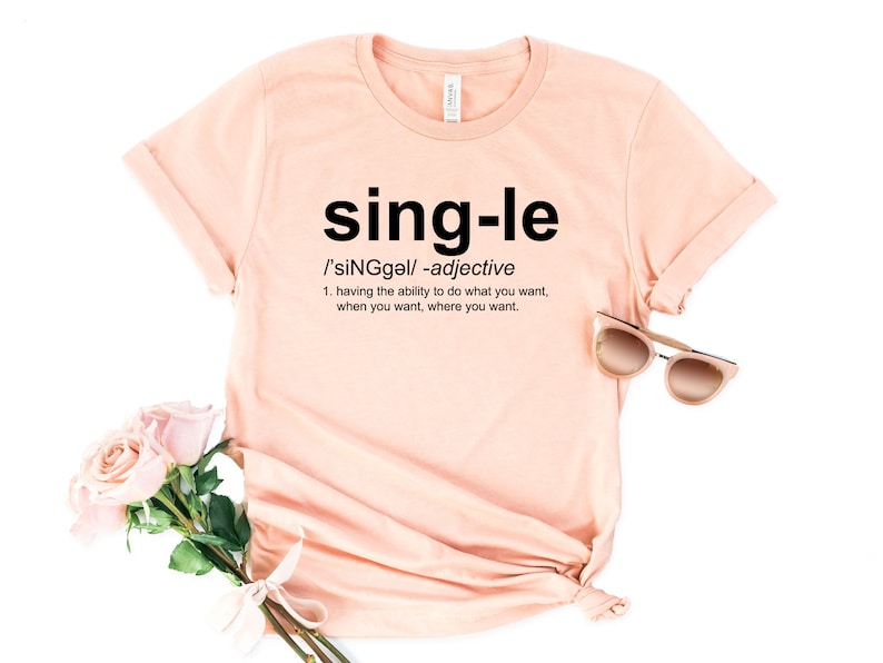 May include: A light peach-colored t-shirt with the word "single" in large black letters. Below the word is the definition of single as an adjective. A pair of sunglasses and a bouquet of pink roses are next to the shirt.