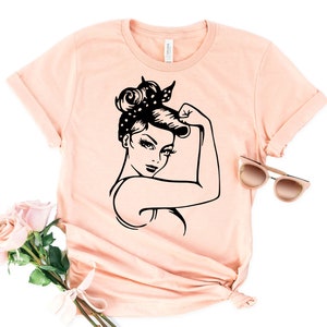 Może przedstawiać: R&oacute;żowy t-shirt z czarno-białym nadrukiem przedstawiającym kobietę z podniesioną pięścią. Kobieta ma na sobie chustkę z białymi kropkami.