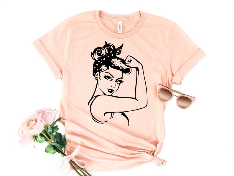 Może przedstawiać: R&oacute;żowy t-shirt z czarno-białym nadrukiem przedstawiającym kobietę z podniesioną pięścią. Kobieta ma na sobie chustkę z białymi kropkami.