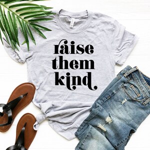 May include: A gray t-shirt with the text "raise them kind" in black lettering. The shirt is folded and laid on a white surface with a pair of brown sandals and blue jeans.