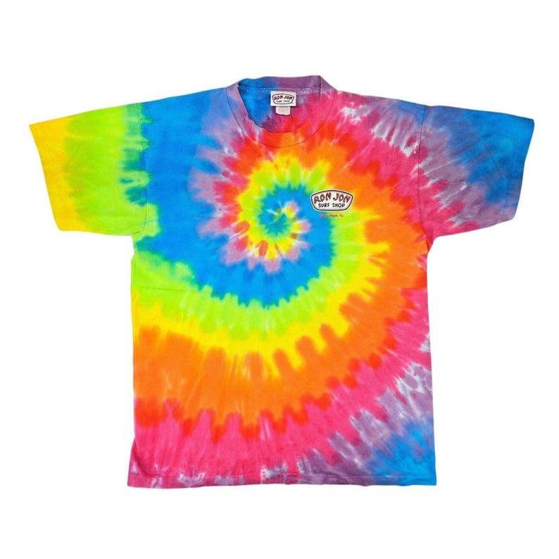 Puede incluir: Una camiseta te&ntilde;ida con un dise&ntilde;o de espiral en colores vibrantes como azul, verde, amarillo, naranja y rosa. La camiseta tiene mangas cortas y cuello redondo. El texto "RON JON SURF SHOP" est&aacute; impreso en la parte delantera.
