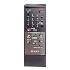 May include: A black Toshiba VC-62 VCR remote control. The remote has a variety of buttons, including power, channel, and play/pause controls. The remote is rectangular and has a silver-colored section with the Toshiba logo.