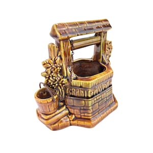 Pode incluir: Um vaso de cerâmica em forma de poço dos desejos com um esmalte castanho e amarelo. O poço apresenta um design semelhante a tijolos, um pequeno balde e o texto "GRANT AVIS". A parte superior tem uma estrutura semelhante a um telhado com uma corrente.