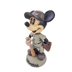Peut inclure: Figurine de Mickey Mouse en uniforme de baseball, tenant une batte et portant une casquette et un gant. La figurine est sur une base avec l'inscription "Disneyland Resort". Les couleurs sont beige, noir, marron et vert.
