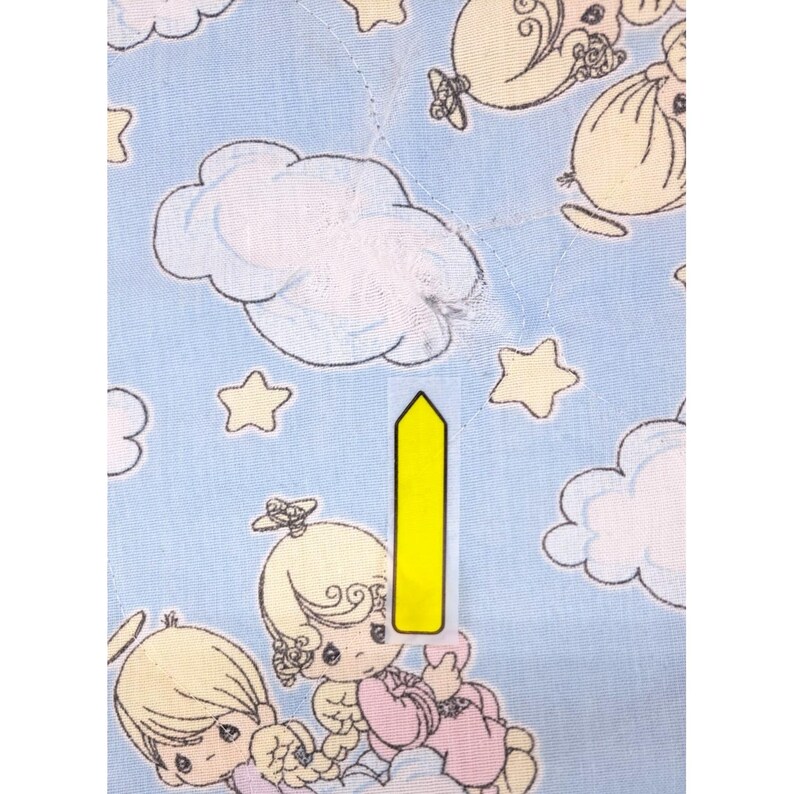 May include: Pale blue fabric featuring a repeating pattern of cartoon angels, clouds, and stars. The angels have blonde hair and wear pink and white clothing. The clouds and stars are yellow. The fabric has a quilted texture.