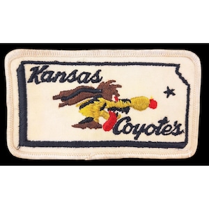 May include: Embroidered patch featuring a cartoon coyote in yellow, brown, and red, with the words "Kansas Coyotes" in black script. The patch has a cream-colored background with a black border and a star graphic.