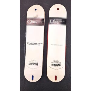 Könnte beinhalten: Zwei Packungen Offray Designer-Bänder. Eine Packung enthält blaues Band und die andere rotes Band. Die Packungen sind weiß mit dem Offray-Logo und dem Text "100% Easy-Care Polyester Crush Resistant Velvet" und "Quality Woven-Edge Ribbons".