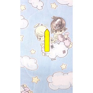 May include: Light blue fabric patterned with clouds and stars, featuring a Precious Moments angel illustration. A quarter coin is placed on the fabric for size comparison. The text "&copy;PMI" is visible.