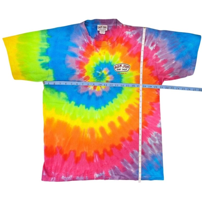 Puede incluir: Una camiseta te&ntilde;ida con un patr&oacute;n de espiral en colores vibrantes como verde, amarillo, naranja, rojo, azul y morado. La camiseta tiene cuello redondo y mangas cortas. El texto "RON JON SURF SHOP" est&aacute; impreso.