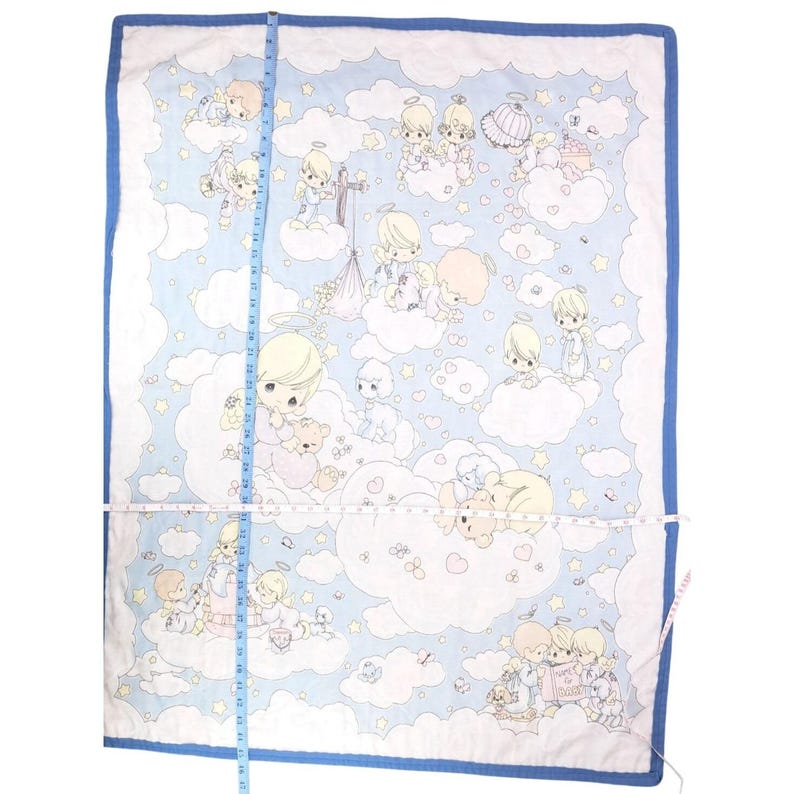 May include: A baby quilt with a light blue backdrop, white clouds, and cherubic figures. The quilt has a blue border and is approximately 76 cm long. The figures are in pastel shades, holding teddy bears and other items.