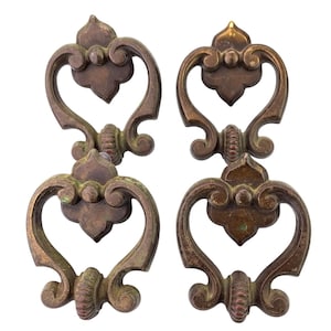 May include: A set of four matching bronze-toned metal drawer pulls featuring a decorative openwork design with a central clover-like shape and a curved, swinging handle. The hardware displays a weathered, aged finish with subtle green patina.