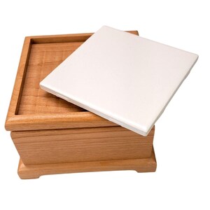 May include: A square wooden box with a light brown natural wood grain finish and a removable white ceramic tile lid. The box features a recessed top edge designed to hold the flat, smooth, square tile securely in place as a cover.