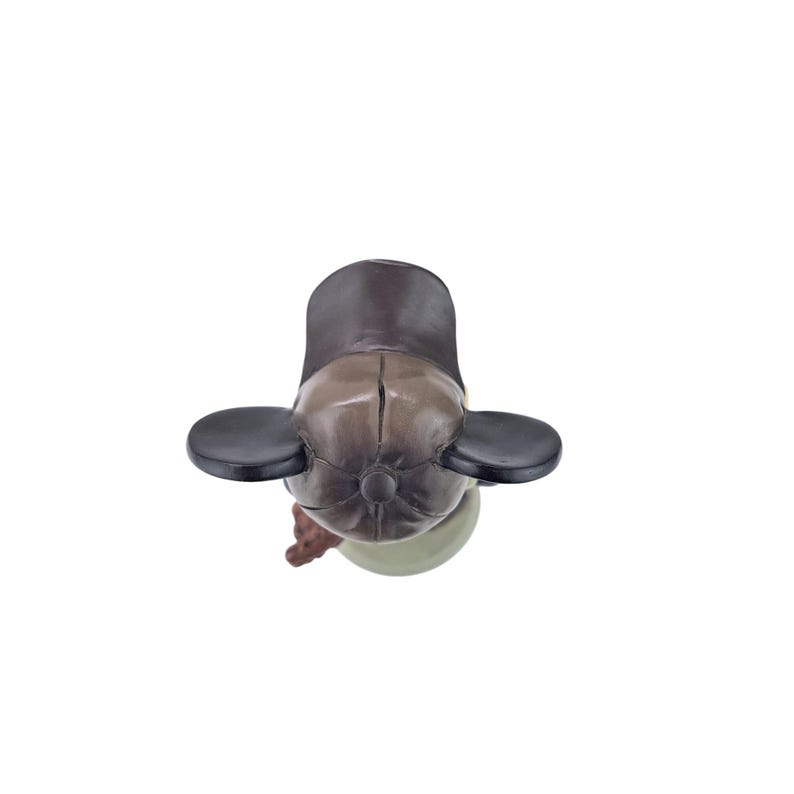 Peut inclure: Figurine d'un personnage portant une casquette marron avec des cache-oreilles noirs. La figurine a un corps vert clair et des bras bruns. La figurine est vue de dessus sur fond blanc.