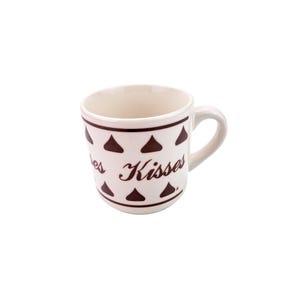 May include: A white ceramic mug with a brown rim and handle. The mug is decorated with repeating brown Hershey's Kisses and the word "Kisses" in a cursive font.