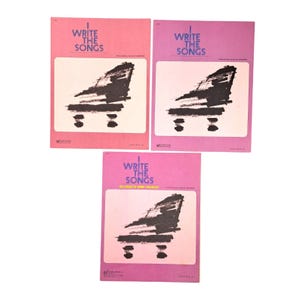 May include: Three sheet music books with the title "I Write The Songs" in white text on a pink and purple background. Each book features a black piano illustration. The books are arranged in a triangular pattern.