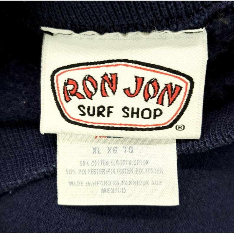 May include: Close-up of a navy blue garment tag. The tag features the red and white "RON JON SURF SHOP" logo. Below the logo are size indicators and fabric content details. The tag reads "MADE IN MEXICO".