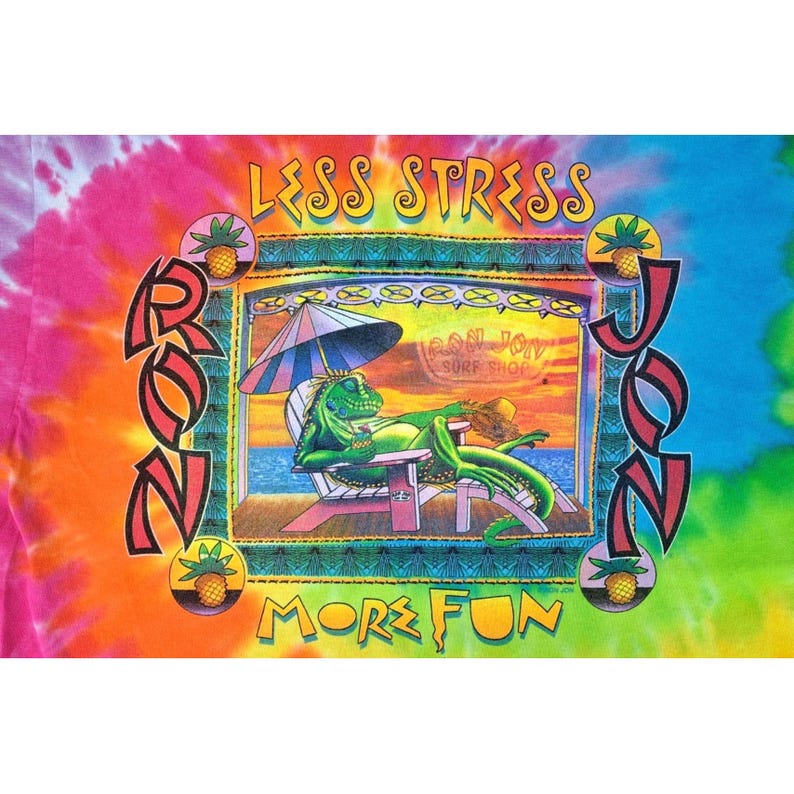 Puede incluir: Camiseta te&ntilde;ida con un dise&ntilde;o de un lagarto verde relaj&aacute;ndose en una silla de playa bajo una sombrilla. El dise&ntilde;o incluye las palabras "LESS STRESS" y "MORE FUN" con el nombre "RON JON" en una fuente estilizada.