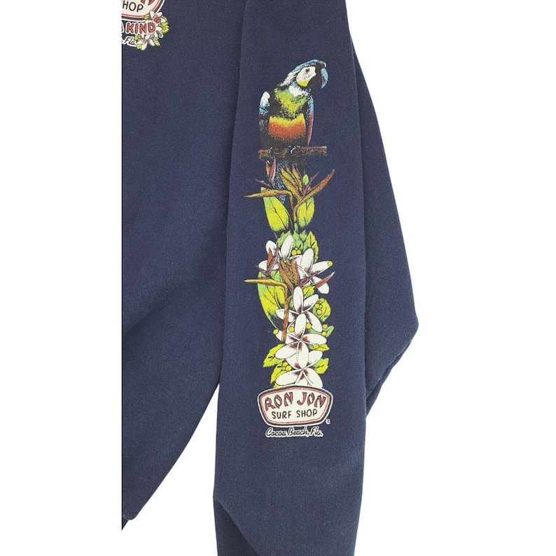 May include: Navy blue sweatshirt with a colorful parrot and floral design on the sleeve. The sleeve also features the text "RON JON SURF SHOP" in a circular logo. The sweatshirt has a crew neck and long sleeves.
