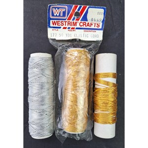 May include: Three spools of metallic cord. One spool is silver, one is gold, and one is a thinner gold cord. The center gold spool is inside a clear plastic Westrim Crafts bag with a header card labeled 177 50 YDS ELASTIC CORD.