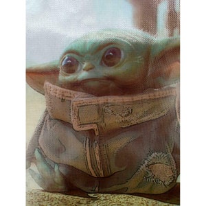 Op de afbeelding: Een close-up van een groen, cartoonachtig personage met grote ogen en een bruin jack. Het personage zit op een lichtbruin oppervlak.