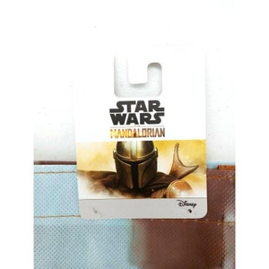 Op de afbeelding: Een wit label met de tekst "Star Wars The Mandalorian" en een afbeelding van de Mandalorian in zijn helm en harnas. Het label heeft een Disney-logo in de rechterbenedenhoek.