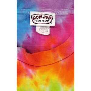 Puede incluir: Una camiseta te&ntilde;ida con un vibrante conjunto de colores, incluyendo azul, morado, naranja y verde. La camiseta tiene una etiqueta con el texto "RON JON SURF SHOP" e instrucciones de lavado. Talla L.