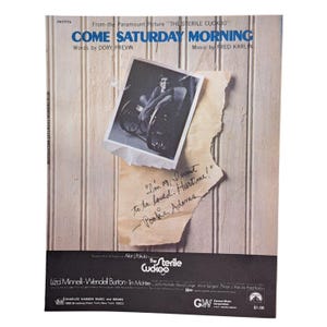 以下が含まれることがあります： 映画「The Sterile Cuckoo」の「Come Saturday Morning」の楽譜カバー。カバーには、白黒写真、引用文が書かれた破れた紙、映画のタイトル、曲名、クレジットなどのテキストが掲載されています。背景は明るい木製のパネルです。