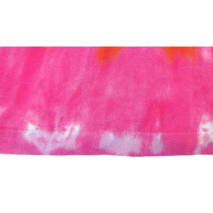 Puede incluir: Primer plano de una tela te&ntilde;ida con un vibrante color rosa como base, acentuada por tonos naranja y blanco. La tela tiene un aspecto suave y texturizado, con costuras visibles a lo largo del borde inferior. El patr&oacute;n tie-dye crea un dise&ntilde;o &uacute;nico y colorido.
