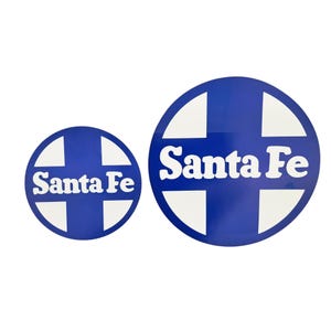May include: Two blue and white Santa Fe stickers. The stickers are circular with a white cross design and the words "Santa Fe" in white. One sticker is larger than the other.
